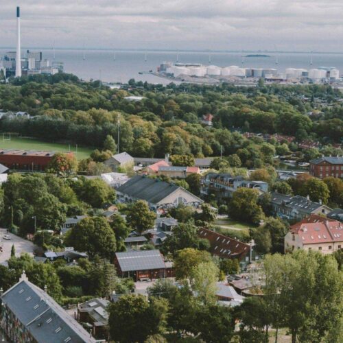 Luftaufnahme einer Stadt mit Grünflächen, Wohnhäusern und Hafenanlage im Hintergrund