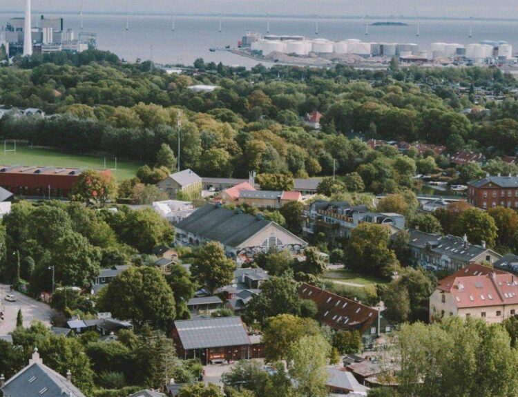 Luftaufnahme einer Stadt mit Grünflächen, Wohnhäusern und Hafenanlage im Hintergrund